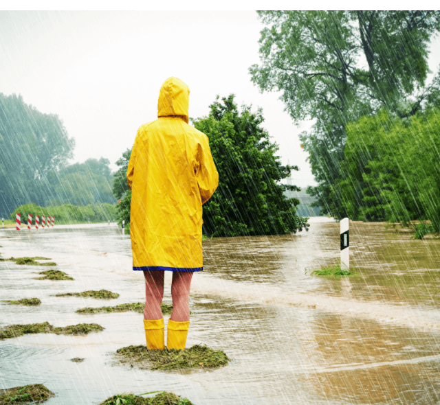 Person im gelben Regenmantel steht auf Grasinsel auf einer überfluteten Straße als Symbolbild für Tipps zu Unwetterschutz der VGH Niedersachsen