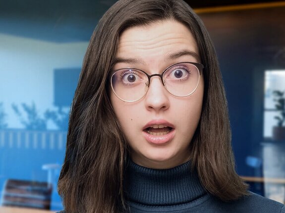 Junge Frau mit Brille und überraschter Miene steht in einem modernen Büroraum.