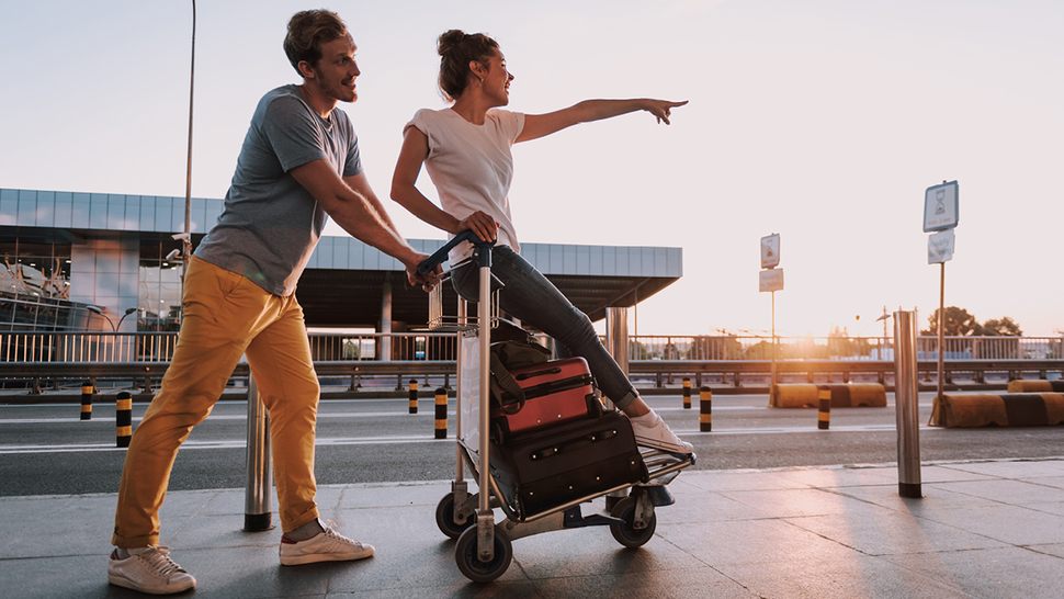 Paar mit Gepäckwagen am Flughafen, der Mann schiebt, während die Frau in die Ferne zeigt. Sonne geht unter.