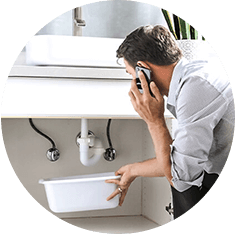 Mann telefoniert und hält eine weiße Schüssel unter ein tropfendes Rohr im Badezimmer.