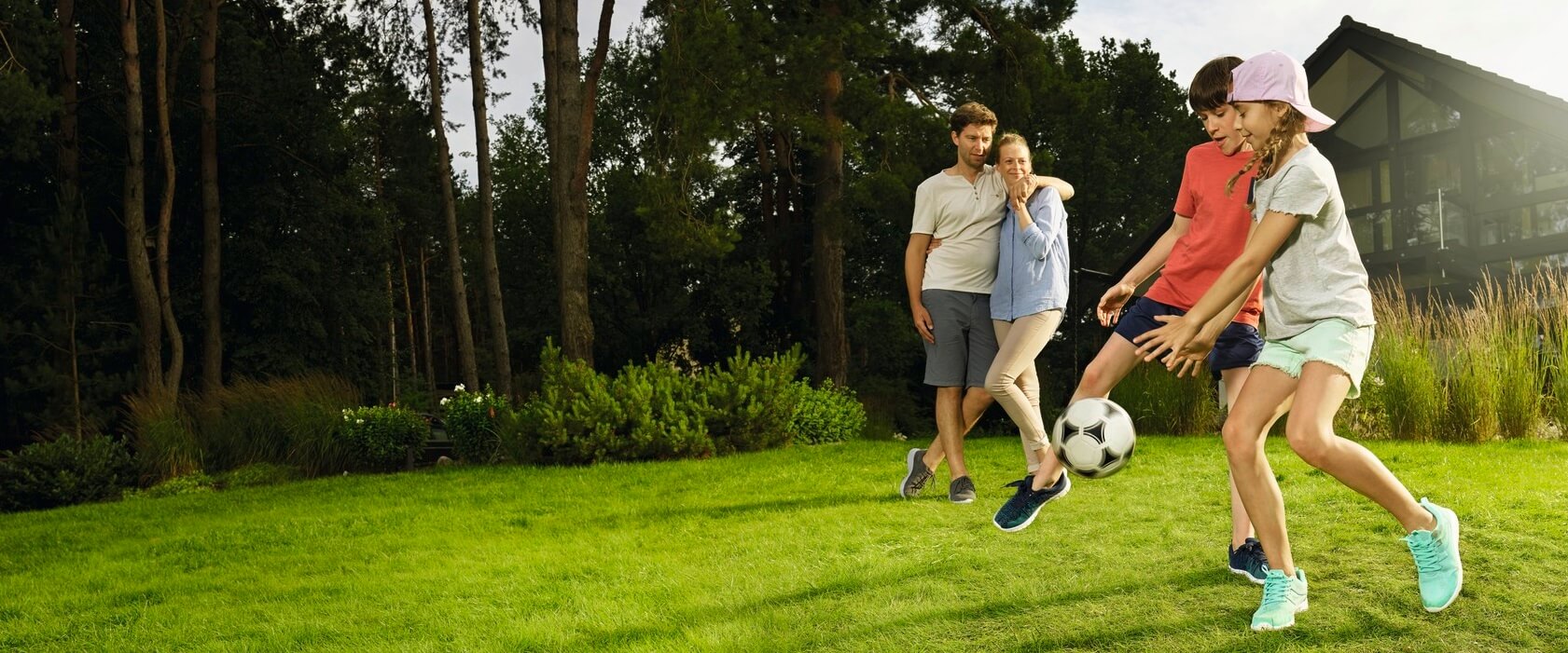 Privathaftpflichtversicherung: Eine vierköpfige Familie im Garten Tochter und Sohn spielen Fußball. Vater und Mutter schauen im Hintergrund zu.