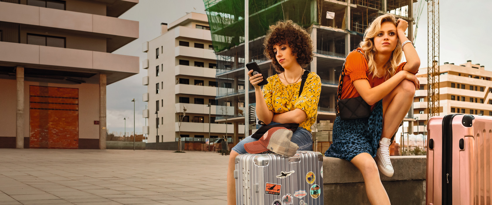 Rechtsschutzversicherung: Zwei Urlauberinnen sitzen enttäuscht mit ihrem Gepäck vor einer Bauruine. Die linke blickt auf Ihr Smartphone.