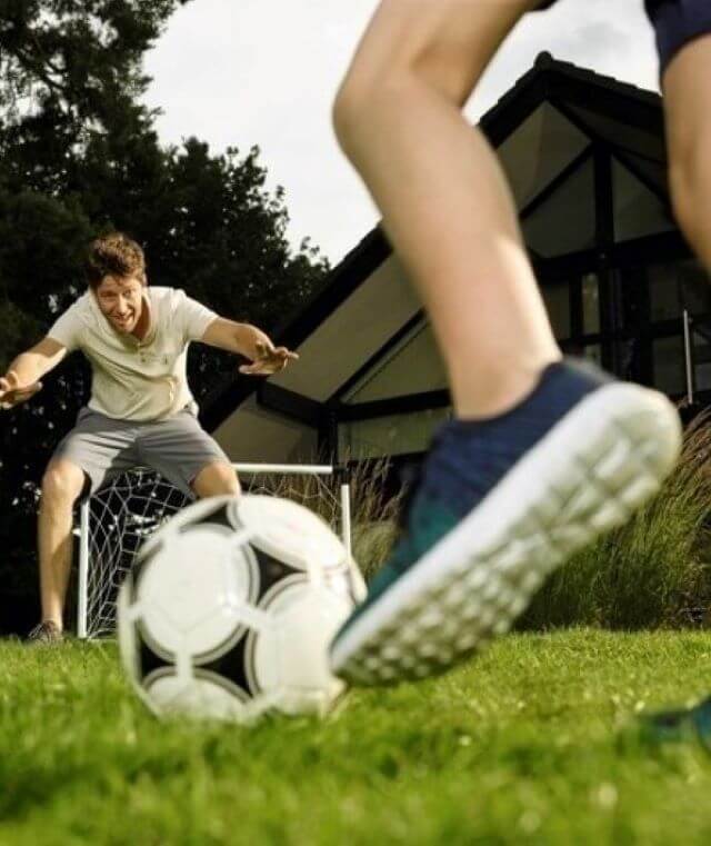 Haftpflichtversicherung: Ein Vater und sein Kind spielen um Garten Fußball. Der Vater steht im Tor. Im Vordergrund ein Kinderfuß und der Ball kurz vor dem Schuss.