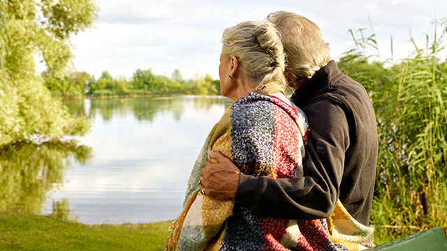 Sterbe­geld­ver­si­che­rung: Ein altes Ehepaar, das bei der VGH versichert ist, stehen am Ufer eines Sees und genießen die Natur. 