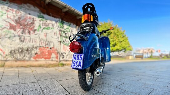 Blick auf ein blaues Moped mit schwarzem Helm, vor einer graffitibedeckten Wand und grünem Baum im Hintergrund.