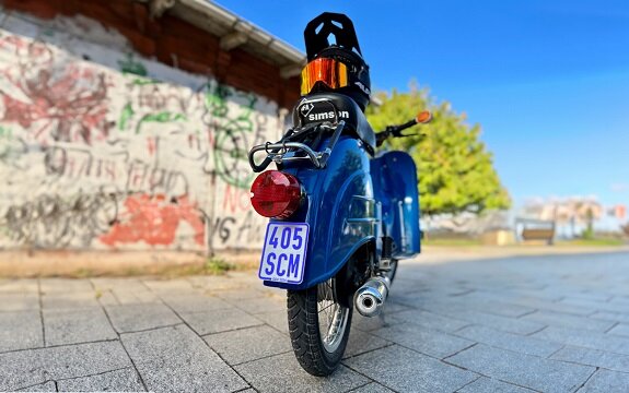 Blick auf ein blaues Moped mit schwarzem Helm, vor einer graffitibedeckten Wand und grünem Baum im Hintergrund.
