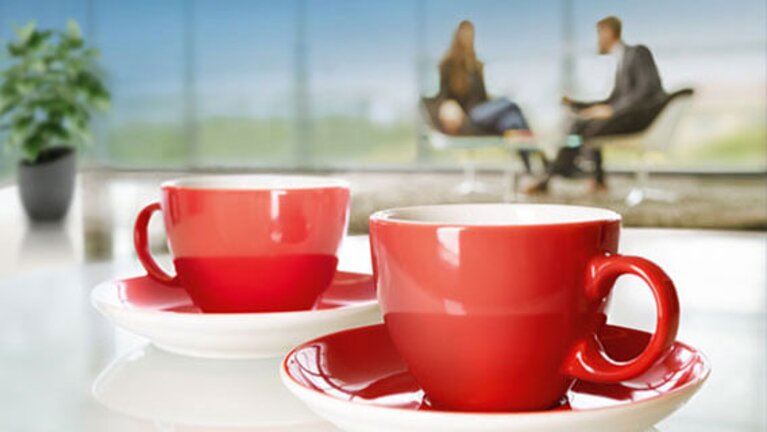 Zwei rote Kaffeetassen stehen auf einem Tisch, im Hintergrund sind zwei Personen im Gespräch zu sehen.