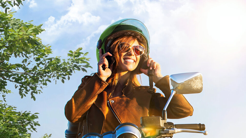 Junge Frau mit Sonnenbrille und Helm lächelt auf einem Motorrad, umgeben von grünen Bäumen und blauem Himmel.