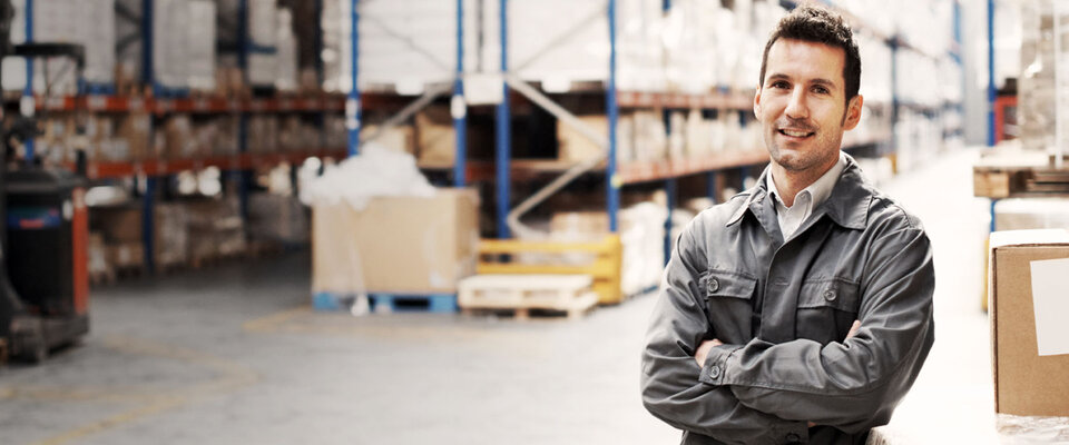 Mann in Arbeitskleidung lächelt in einem Lager mit Regalen voller Kartons im Hintergrund.