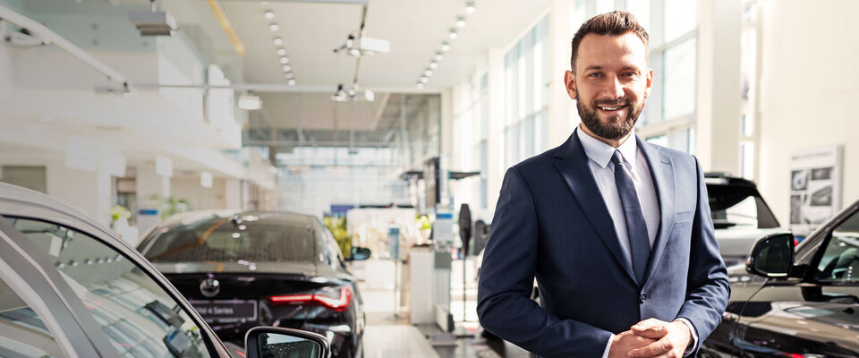 Mann im Anzug steht in einem Autohaus, umgeben von mehreren Fahrzeugen, und lächelt freundlich.