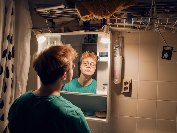Junger Mensch mit einem grünen Shirt steht vor einem Spiegel im Badezimmer und betrachtet sein Spiegelbild.