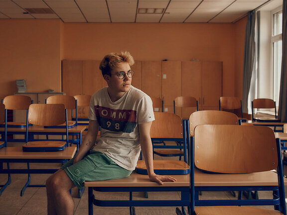 Junger Mann mit Brille sitzt auf einem Schultisch in einem leeren Klassenraum mit orangefarbenen Wänden.