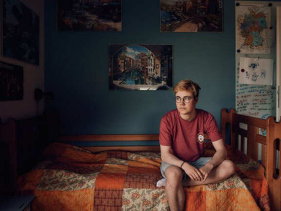 Junger Mann mit Brille sitzt nachdenklich auf einem Bett in einem bunten, gemütlichen Zimmer mit Bildern an der Wand.