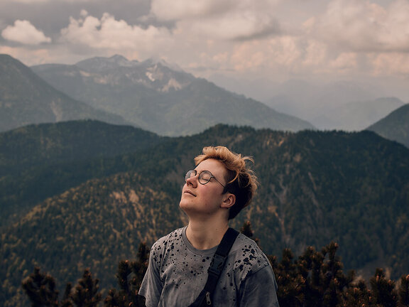 Junger Mensch mit Brille genießt die Aussicht auf eine bergige Landschaft unter einem bewölkten Himmel.