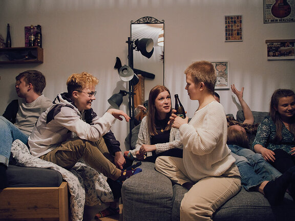 Sechs junge Menschen in einem gemütlichen Raum sitzen auf Sofas, interagieren und lachen miteinander. Gemütliche Atmosphäre.