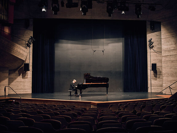 Ein Pianist sitzt an einem Flügel auf einer leeren Bühne mit Dunkelblauem Vorhang und Zuschauerplätzen im Vordergrund.