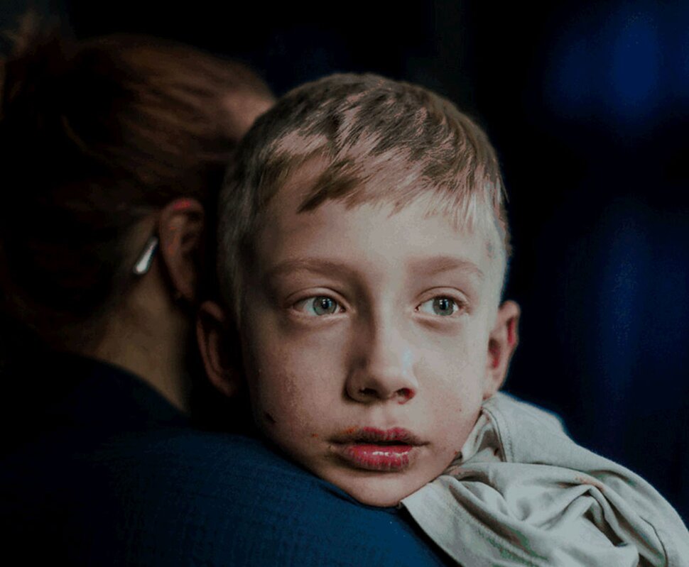 Ein junger Junge mit blonden Haaren und grünen Augen blickt nachdenklich von der Schulter einer Person weg.