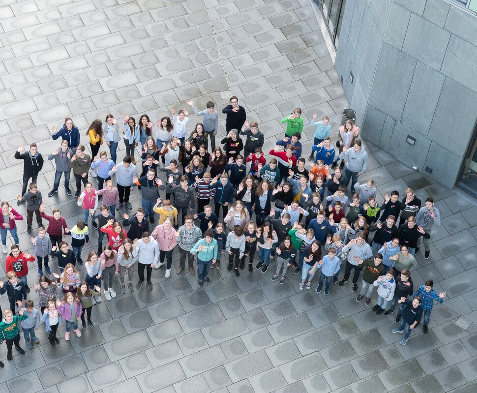 Große Gruppe von Menschen, die auf einem Platz stehen und in die Kamera winken, umgeben von modernen Gebäuden.