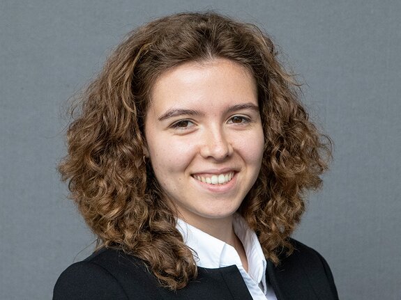 Junge Frau mit lockigem, braunem Haar lächelt, trägt einen schwarzen Blazer und ein weißes Hemd vor grauem Hintergrund.