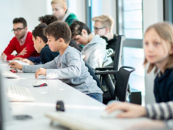 Gruppe junger Menschen sitzt an PC-Arbeitsplätzen und konzentriert sich auf ihre Bildschirme.