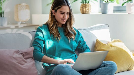 Junge Frau sitzt auf einer Couch, nutzt einen Laptop, umgeben von Kissen und einer hellen, gemütlichen Wohnatmosphäre.
