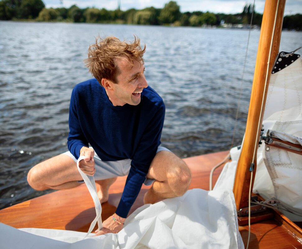 Mann mit einem Rucksack und einer Jacke steht an einem Yachthafen neben einem Segelboot. Sonne scheint am Abend.