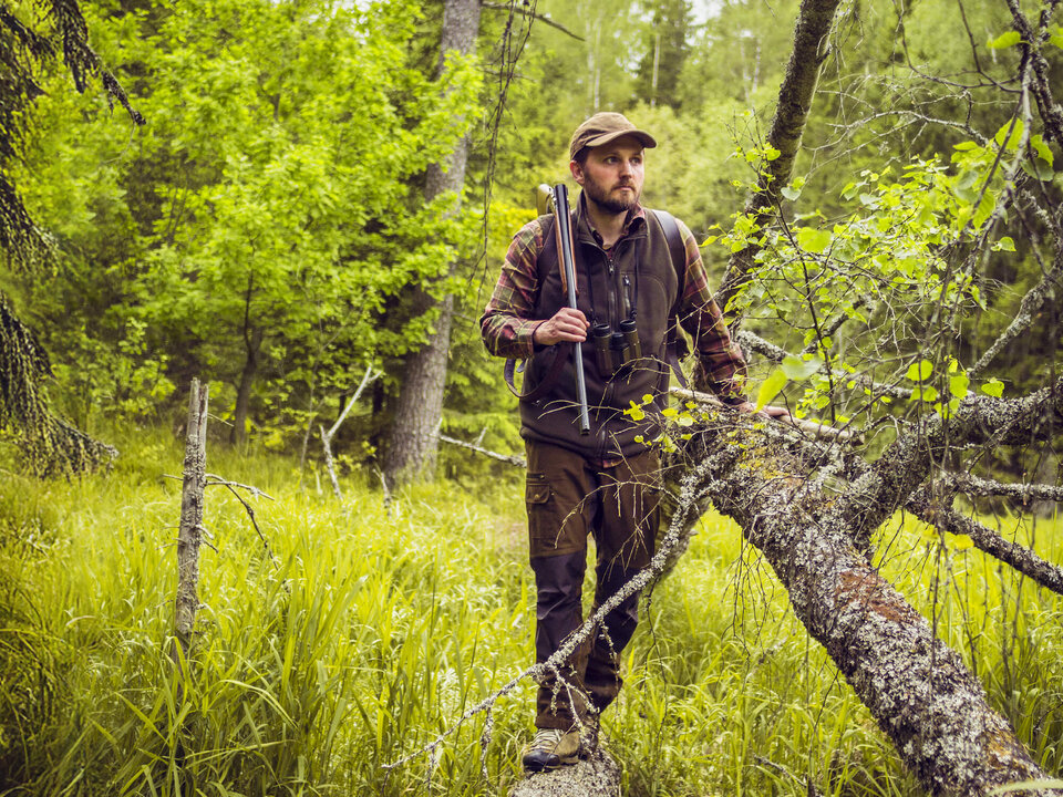 Mann im Wald, bekleidet mit Jagdausrüstung, steht auf einem umgefallenen Baumstamm und hält ein Gewehr in der Hand.