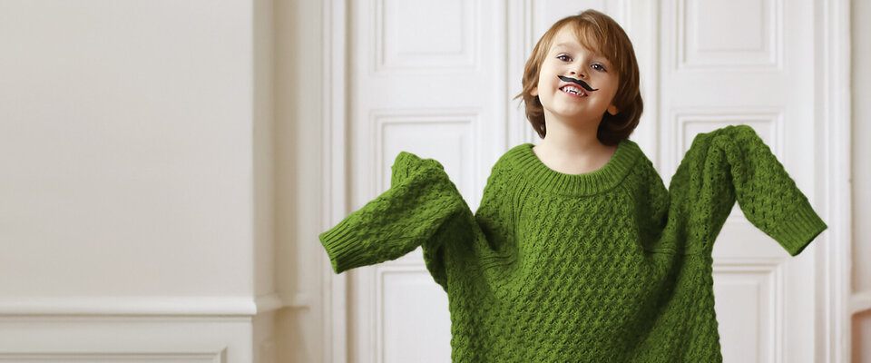 Ein fröhliches Kind in einem oversized grünen Pullover mit einer Spielzeugmustache auf dem Gesicht, das lacht.