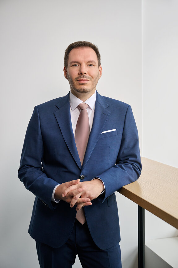 Mann in einem blauen Anzug mit rosa Krawatte, lächelt und steht an einem Holztisch vor einer hellen Wand.