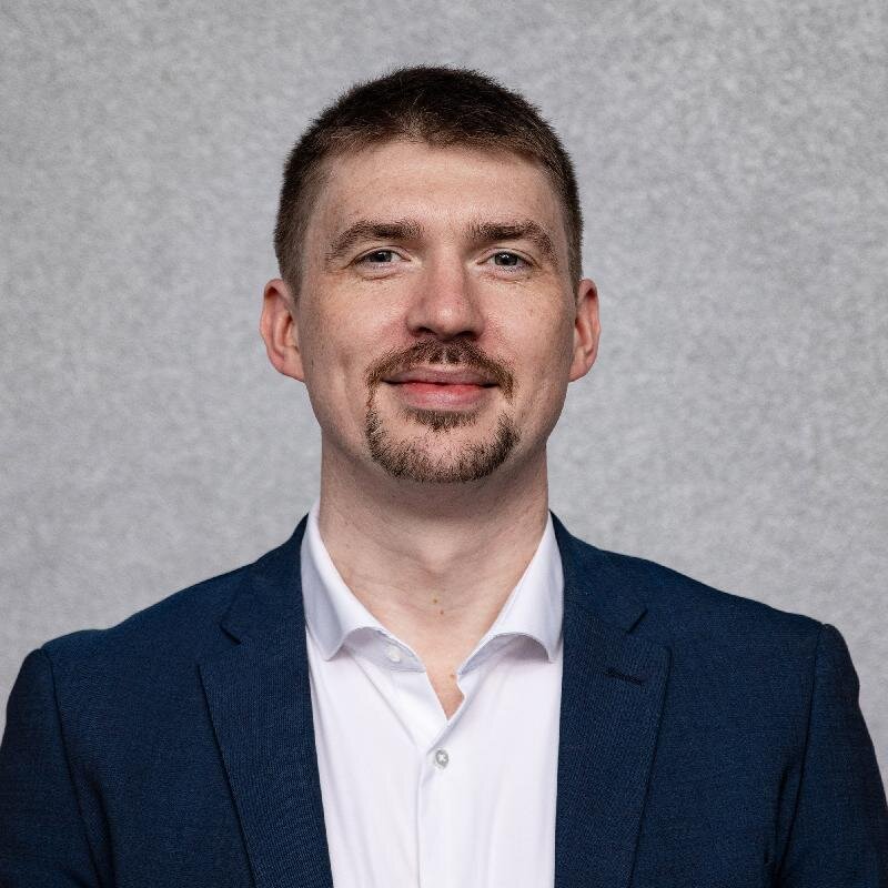 Junger Mann mit braunem Haar und Bart, weißem Hemd und blauem Sakko lächelt vor neutralem Hintergrund.