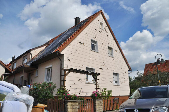 Die Fassadenplatten eines zweigeschossigen Wohnhauses mit Giebeldach sind nach Hageleinschlägen stark beschädigt und weisen große Löcher auf.