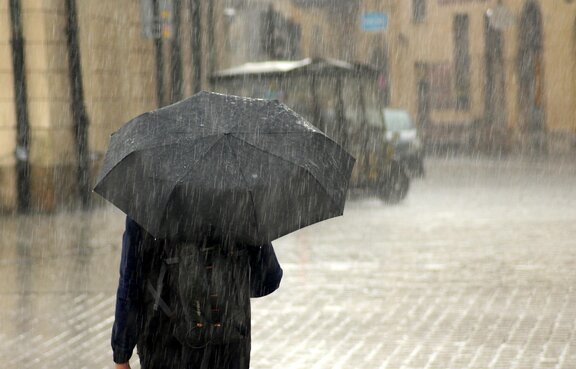 Eine Person steht mit dem Rücken zum Fotografen auf einem städtischen Platz. Es regnet stark, die Person hält einen Regenschirm schützend über sich. Im Hintergrund sind unscharf Häuserzeilen und Fahrzeuge zu sehen.