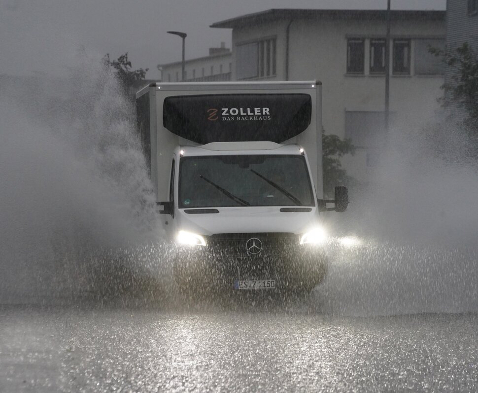 Ein Kleintransporter fährt bei prasselndem Starkregen über eine Straße. Das auf dem Asphalt stehende Wasser spritzt links und rechts vom Fahrzeug hoch auf.