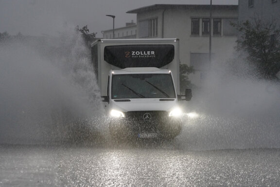 Ein Kleintransporter fährt bei prasselndem Starkregen über eine Straße. Das auf dem Asphalt stehende Wasser spritzt links und rechts vom Fahrzeug hoch auf.