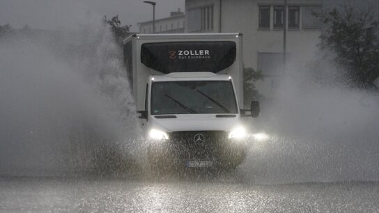 Ein Kleintransporter fährt bei prasselndem Starkregen über eine Straße. Das auf dem Asphalt stehende Wasser spritzt links und rechts vom Fahrzeug hoch auf.
