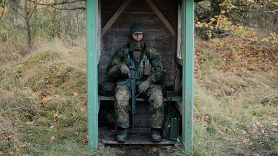 Ein Soldat in Tarnumiform sitzt mit Gewehr in einem Wachhäuschen, das mitten im Wald steht.