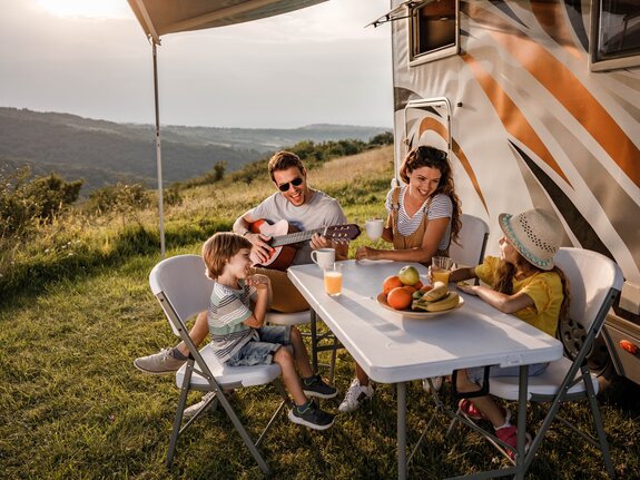 Familie sitzt entspannt am Tisch vor einem Wohnmobil, genießt Snacks und Musik in der Natur.
