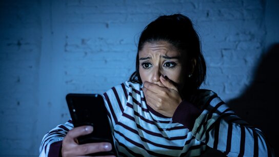Eine junge Frau schaut besorgt auf ihr Smartphone und hält sich besorgt die Hand vor den Mund. Gedämpftes Licht von hinten.