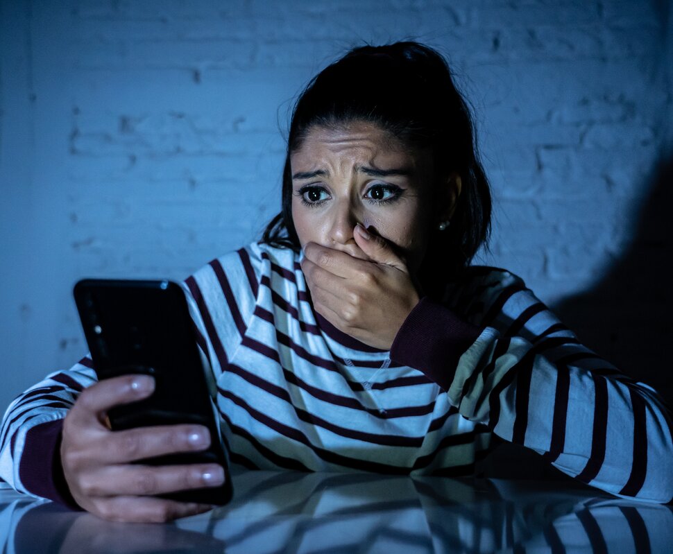 Eine junge Frau schaut besorgt auf ihr Smartphone und hält sich besorgt die Hand vor den Mund. Gedämpftes Licht von hinten.