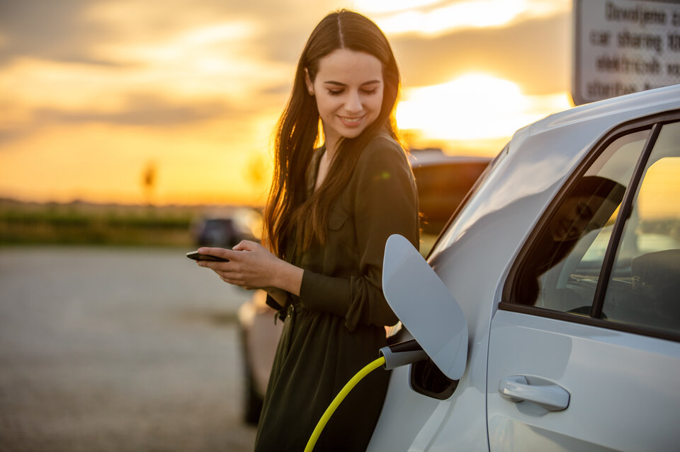 Junge Frau in einem Kleid lädt ein Elektroauto auf und hält ein Smartphone in der Hand, während die Sonne untergeht.