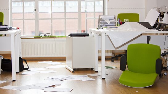 Unordentliches Büro mit verstreuten Papieren, leeren Stühlen und grünem Bürostuhl, der umgekippt ist.