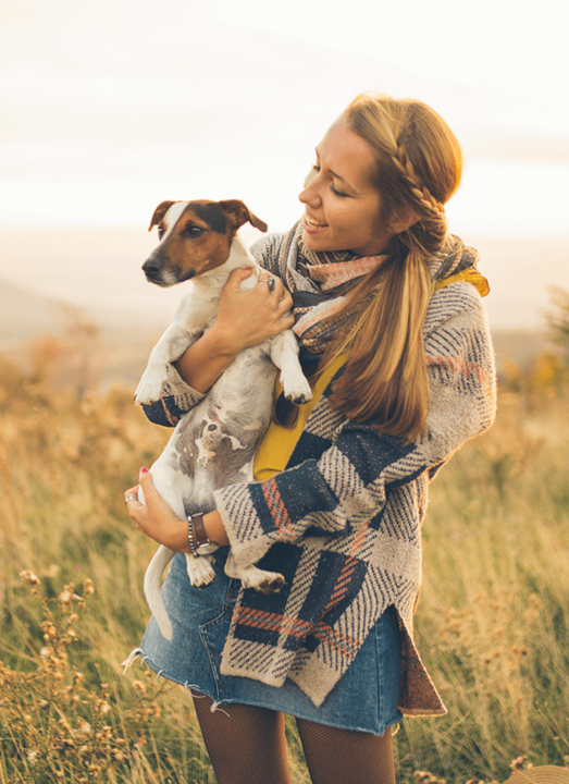 Frau mit Zopf hält einen kleinen Hund. Sie lächelt und steht in einem grünen Feld bei Sonnenuntergang.