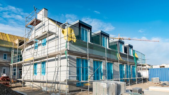 Neubau mit Gerüst, blauen Fenstern und Baustellenmaterialien, umgeben von einem klaren Himmel.
