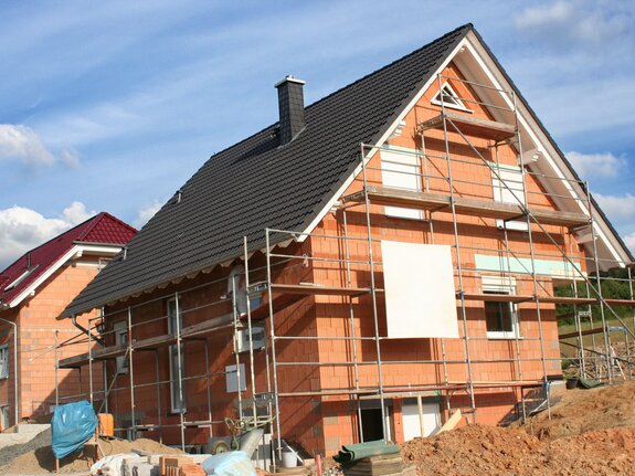 Neubau eines Hauses mit Gerüst, teils mit Fassade verkleidet, im Hintergrund weitere Bauarbeiten und blaue Himmel.