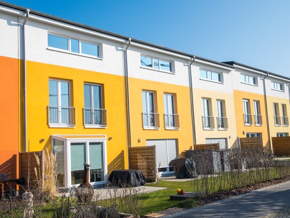 Reihenhäuser mit bunten Fassaden in Orange und Gelb unter klarem, blauem Himmel. Gärten und Terrassen im Vordergrund.