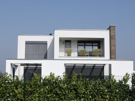 Modernes Haus mit klaren Linien, großen Fenstern und Balkon, umgeben von einer grünen Hecke und blauen Himmel.
