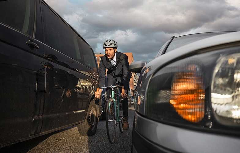 Mann im Anzug und Helm fährt mit dem Fahrrad durch enge Lücke zwischen zwei Autos unter bewölktem Himmel.