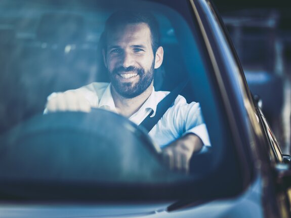 Lächelnder Mann mit Vollbart sitzt am Steuer eines Autos und hält das Lenkrad, Blick nach vorne gerichtet.