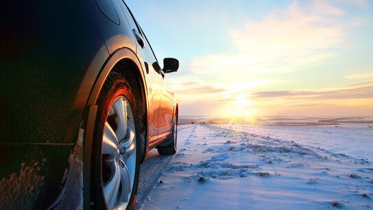 Schwarzes Auto auf einem verschneiten Weg, im Hintergrund strahlender Sonnenuntergang über einer frostigen Landschaft.