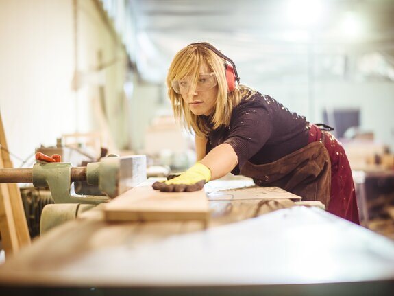 Frau in einer Werkstatt, die mit Handschuhen ein Holzbrett bearbeitet und dabei Gehörschutz und Schutzbrille trägt.
